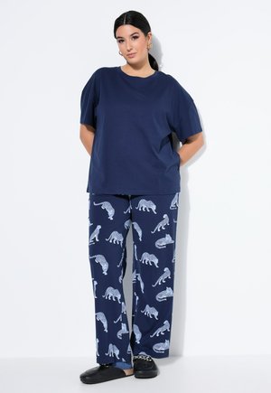 Vrouw staand met handen achter haar rug, gekleed in een donkerblauw t-shirt en donkerblauwe broek met witte luipaardprint en zwarte instapschoenen.