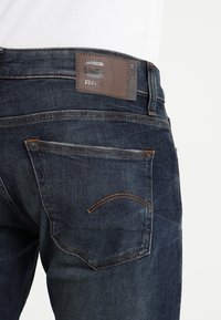 Mörka denimjeans med en brun läderlapp på midjan, synliga bakfickor och kontrasterande oranga sömdetaljer.