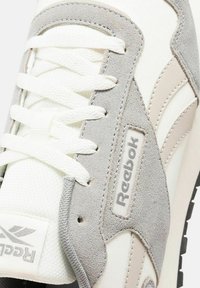 Zapatilla blanca con acentos de ante gris y malla texturizada. Presenta cordones blancos planos y un logo de Reebok en el costado. Suela negra.