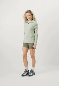 Μακρυμάνικος, ανοιχτό πράσινο πόλο πουφάν με κοπή στο ένα τέταρτο του λαιμού, συνδυασμένος με ελαιόχρωμα σορτς και γκρί παπούτσια πεζοπορίας με άσπρες λεπτομέρειες.