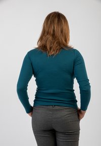 Maglietta fitted a maniche lunghe color teal, con texture a coste e scollatura rotonda. Abbinata a pantaloni grigi, mostrata da dietro. Dettagli minimal.