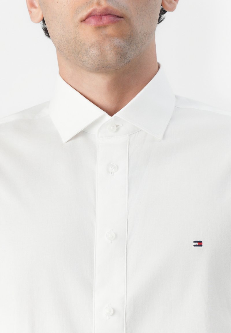 Chemise blanche à boutons avec un col pointu. Présente un petit logo brodé en marine, rouge et blanc sur la poitrine gauche. Texture de tissu douce.