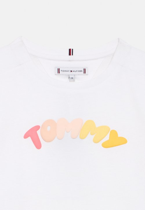 T-shirt blanc à col rond, avec le texte coloré "TOMMY" en lettres arrondies sur la poitrine et une étiquette de marque Tommy Hilfiger à l'intérieur du col.