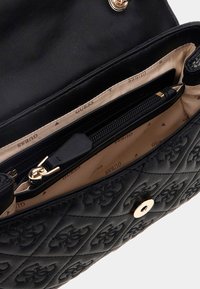 Schwarze gesteppte Handtasche mit goldenen Metallteilen, Innenfach mit Reißverschluss und beigem Futter mit dezentem Logo-Muster. Mit einem Druckknopf verschlossen.