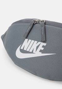 Pelēka auduma fanny pack ar gludu tekstūru, kas rotāta ar izteiktu baltu Nike logo un diviem metāla rāvējslēdzējiem. Kompakta, izliekta forma.