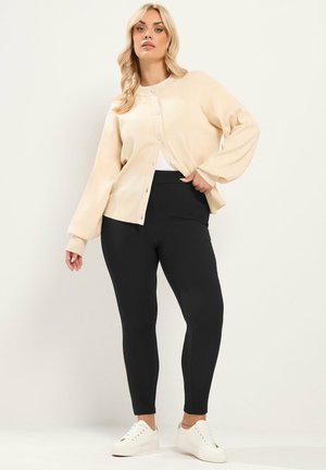 Blondine vrouw draagt een beige gebuttonde cardigan, zwarte aansluitende broek en witte sneakers en poseert tegen een effen lichte achtergrond.