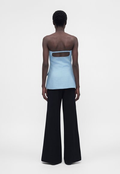 Άτομο που φοράει αμάνικη γαλάζια μπλούζα με ορθογώνιο άνοιγμα στο πάνω μέρος της πλάτης και φαρδιά μαύρα παντελόνια, στέκεται σε απλό φόντο.