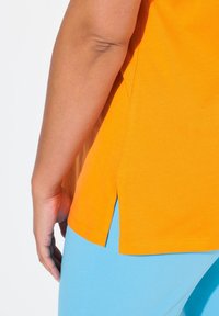 Orange bomulds-t-shirt med korte ærmer, sideslids og en glat tekstur, parret med lyseblå bukser.