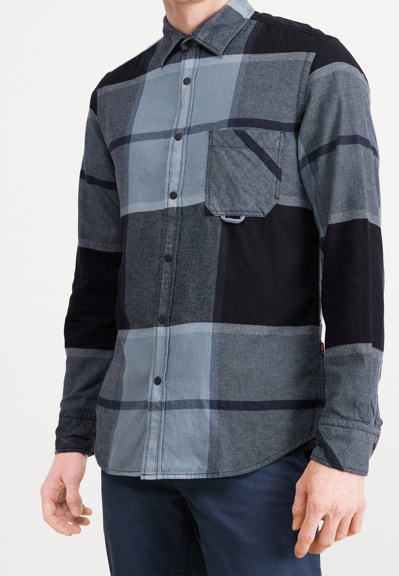 Homme portant une chemise à manches longues à boutons avec un grand motif à carreaux noir, gris et bleu et une poche poitrine, associée à un pantalon foncé.