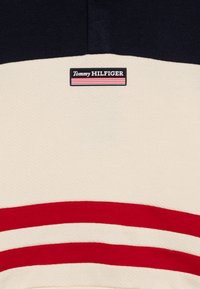 Polo-skjorte med marineblå øvre seksjon, kremfarget nedre seksjon og røde striper. Har et rektangulært logo med tekstene "Tommy HILFIGER". Teksturert stoff.