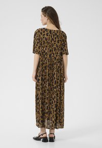 Robe maxi brune avec un imprimé léopard noir. Elle présente des manches courtes, une coupe ample et des détails plissés à la taille. Chaussures noires avec des talons épais.