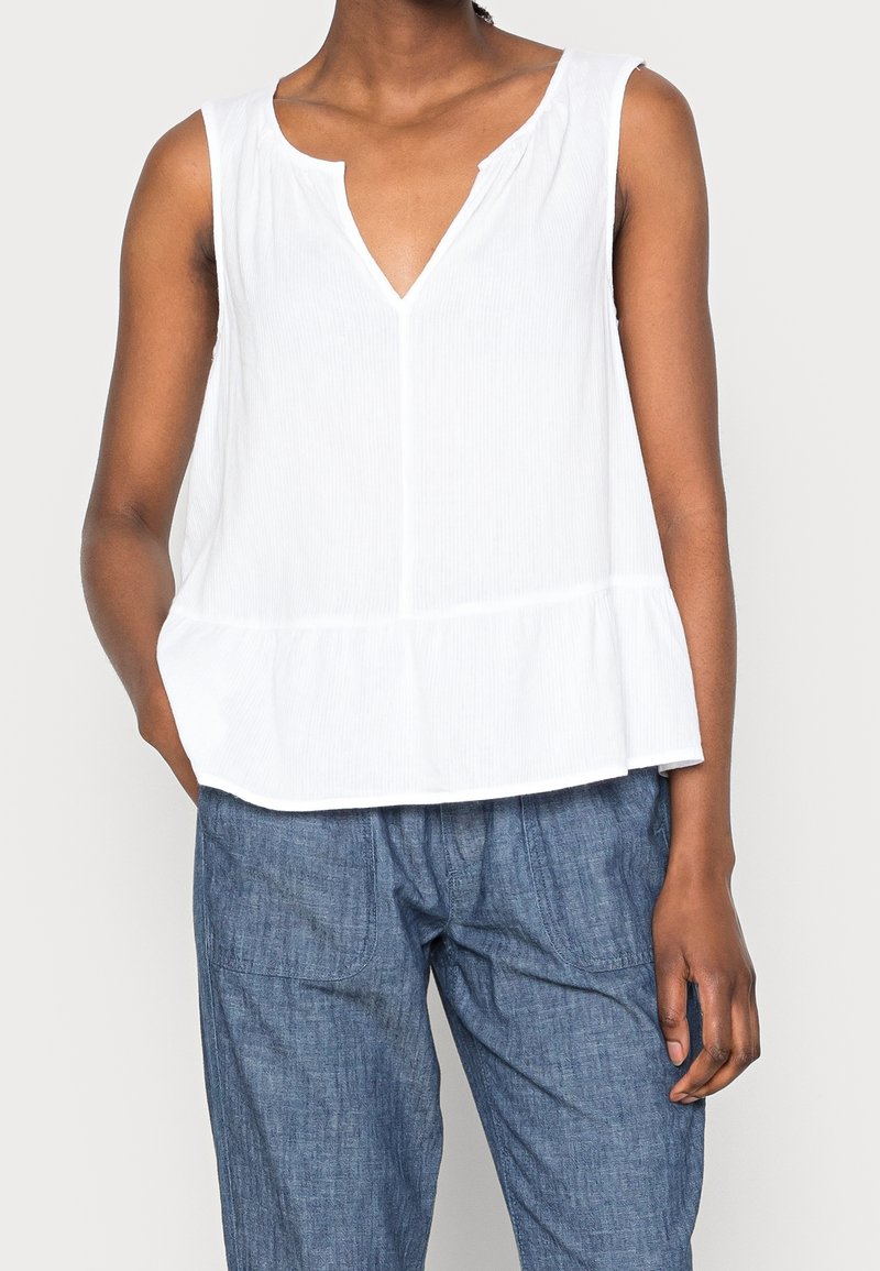 Personne portant un haut blanc sans manches avec une encolure fendue et un jean bleu, debout avec une main dans la poche sur un fond uni.