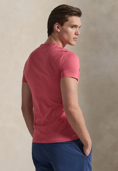 Εφαρμοστό κοντομάνικο t-shirt σε κοραλί ροζ βαμβάκι, λεία υφή, λαιμόκοψη crew, εμφανίζεται από πίσω με μπλε σορτς και ουδέτερο φόντο.