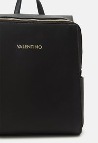 Černý kožený batoh s texturovaným povrchem, zlatým logem „Valentino“, horním zapínáním na zip a dvojitými uchy pro nošení.