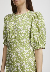 Robe florale verte avec des manches bouffantes courtes, une taille froncée et un motif de fleurs blanches avec des accents jaunes sur un tissu texturé.