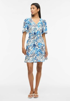 Kobieta stojąca, ubrana w krótką niebiesko-białą sukienkę w kwiaty z falbanowymi rękawami i białe sandały na białym tle.