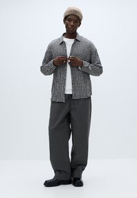 Sort-hvid ternet skjorte med knapper, lagdelt over en hvid t-shirt. Grå bukser med vide ben og sorte støvler. Modellen har en beige strikket hue på.