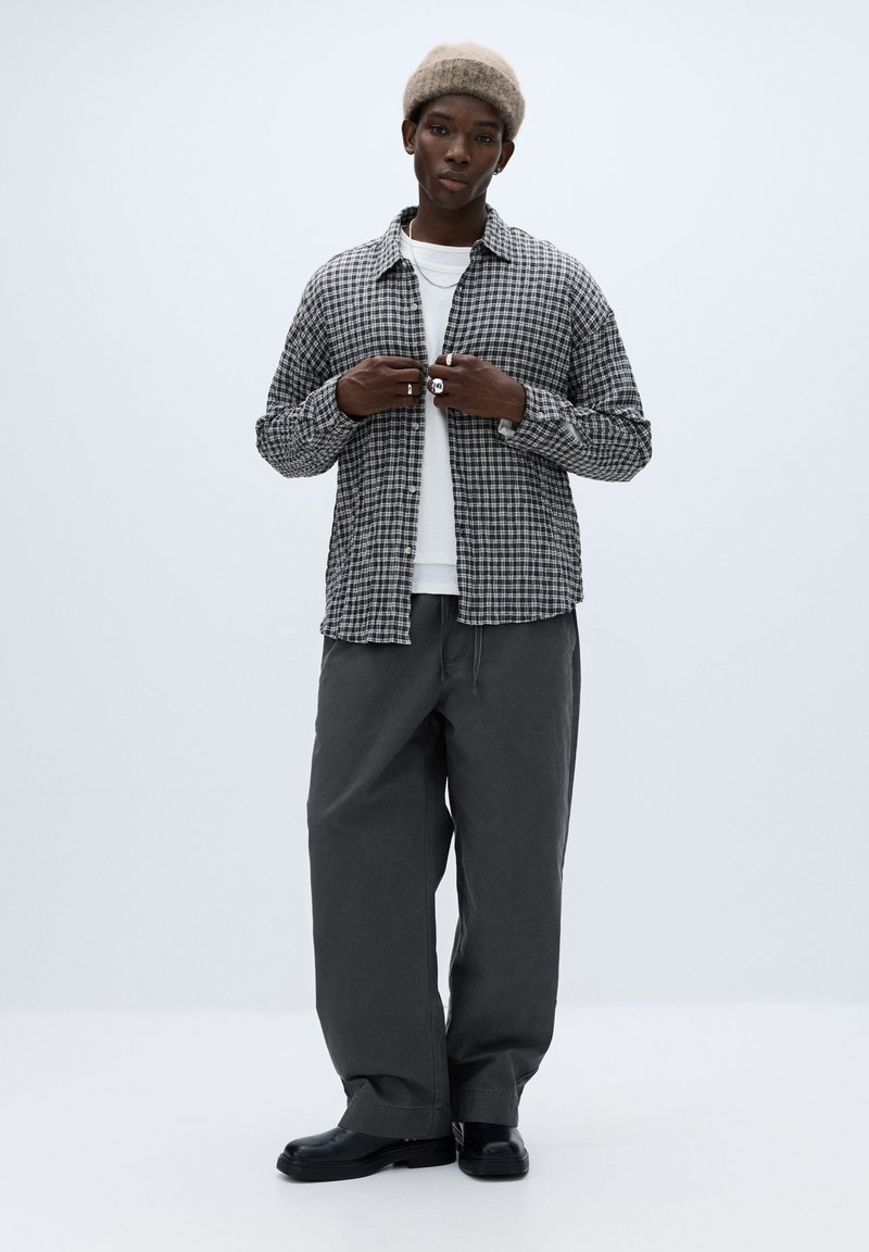 Sort-hvid ternet skjorte med knapper, lagdelt over en hvid t-shirt. Grå bukser med vide ben og sorte støvler. Modellen har en beige strikket hue på.