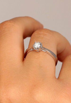 Silberner Ring mit einem zentralen runden, klaren Diamanten, der eine glatte Band und ein minimalistisches Design aufweist, und auf einer Hand mit strukturierter Haut liegt.