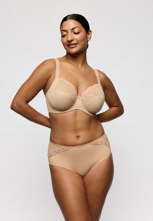 Vrouw die een beige beha met kanten rand en bijpassend hooggetailleerd ondergoed draagt, staat met haar handen achter haar rug en kijkt naar beneden met een lichte glimlach.