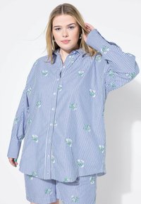 Camicia a righe blu e bianca, con ricami floreali verdi e bianchi. Vestibilità ampia con maniche lunghe e orlo curvo.