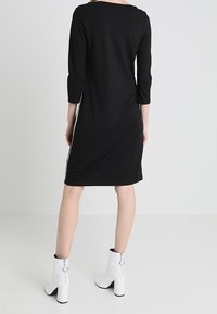 Svart knä-lång klänning med trekvartslånga ärmar, sidostripes och rundad halsringning, kombinerad med vita klackade ankelskor.