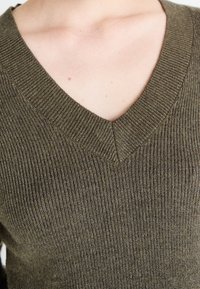 Maglione a coste color verde oliva con scollatura a V, caratterizzato da una texture morbida e da una leggera lucentezza. Inquadratura ravvicinata del colletto e dei dettagli del tessuto.