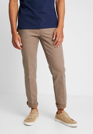 Mężczyzna w beżowych, dopasowanych spodniach podwiniętych przy nogawkach, w połączeniu z brązowymi zamszowymi trampkami i granatową koszulą.