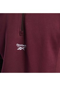 Gros plan du tissu bordeaux d'un sweat-shirt Reebok avec une fermeture éclair noire et un logo Reebok brodé en blanc sous la fermeture éclair.