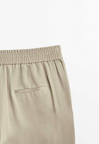 Beżowe spodnie z elastycznym paskiem w talii i jedną kieszenią na plecach, wyeksponowane na białym tle.