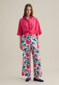 Camicia button-up rosa brillante annodata in vita, abbinata a pantaloni floreali a gamba larga con colori e fantasie vivaci. Scarpe basse blu completano il look.