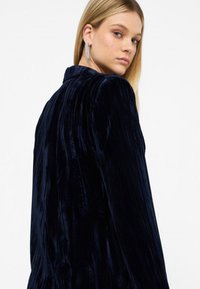 Chaqueta de terciopelo azul marino con textura acanalada vertical, mangas largas y cuello puntiagudo, que presenta un acabado de tela suave y lujoso.