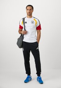 Roupa desportiva Adidas, incluindo uma t-shirt branca de mangas curtas com mangas vermelhas e detalhes em preto, combinada com calças de treino pretas e sapatilhas azuis.