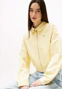 Geel cropped jasje met een kraag en een rits aan de voorkant, met een klein logo op de borst. Model draagt het met lichtblauwe denim jeans.