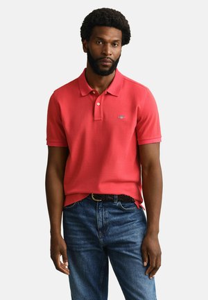 Uomo con acconciatura afro, indossa una polo rossa a maniche corte e jeans blu, in piedi davanti a uno sfondo semplice.