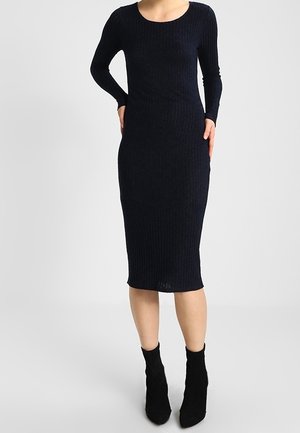 Kobieta nosząca dopasowaną, długą, prążkowaną, ciemnogranatową sukienkę midi z czarnymi botkami, stojąca na jednolitym jasnoszarym tle.