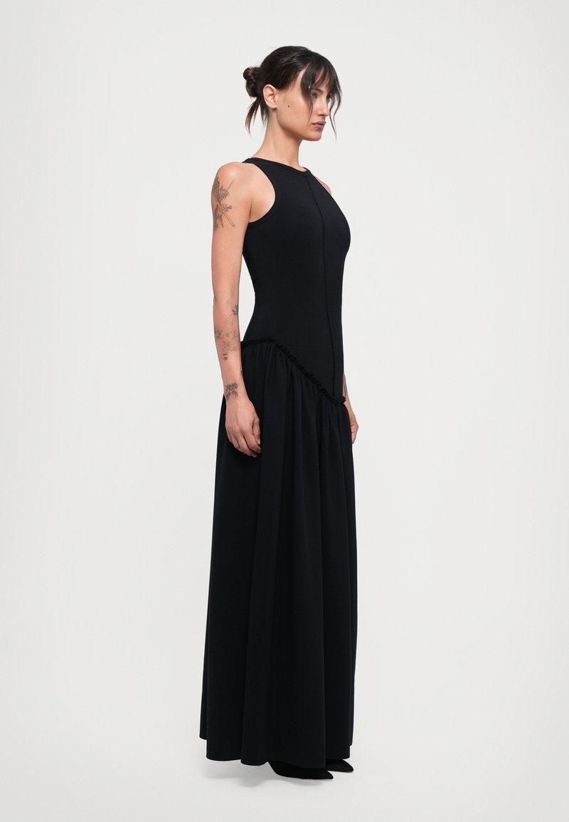 Woman with tattoos wearing a sleeveless black floor-length dress with gathered detail around the hips, standing sideways on a white background.