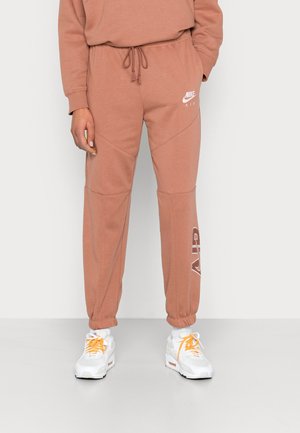 Osoba ubrana w brązowe spodnie dresowe Nike Air oraz białe sneakersy z pomarańczowymi sznurowadłami, stojąca na jednolitym białym tle.