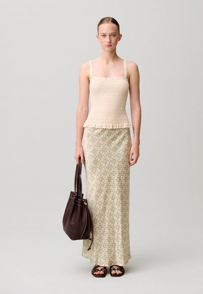 Donna che indossa un top smock color beige, gonna lunga a fantasia, sandali neri, che tiene una borsa in pelle marrone scuro, in piedi su uno sfondo semplice.