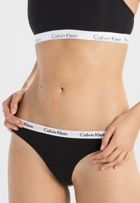 Femme portant un ensemble de sous-vêtements noirs Calvin Klein, ajustant le côté du slip, montrant son ventre et la marque sur la ceinture.