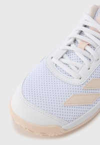 Zapatilla deportiva blanca con parte superior de malla, detalles en rosa y suela de goma. Cuenta con cordones planos y dos franjas laterales texturizadas.