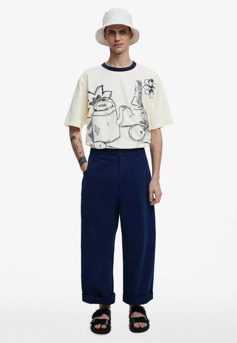 Cappello a secchiello bianco, camicia gialla oversize con disegni neri, pantaloni blu navy e sandali neri. Presenta una vestibilità rilassata e maniche arrotolate.
