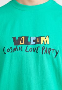 Green cotton T-shirt featuring a bold graphic design with "VOLCOM" in black, yellow, and blue text, and "COSMIC LOVE PARTY" in black.