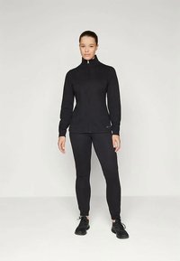 Woman standing, wearing a black zip-up jacket, matching black pants, and black sneakers, against a plain light background.