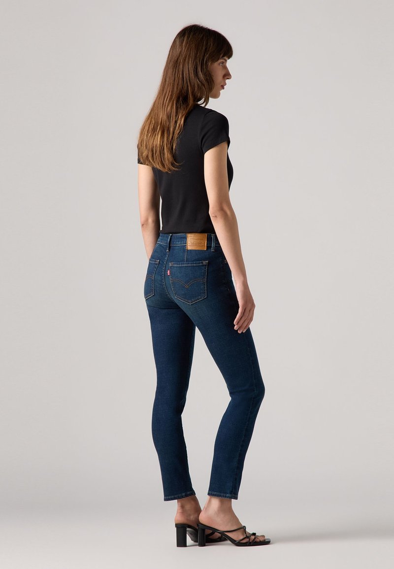 Dünne, dunkelblaue Jeans mit einem Lederpatch am hinteren Bund, kombiniert mit einem eng anliegenden schwarzen Oberteil und schwarzen Riemchen-Heels.