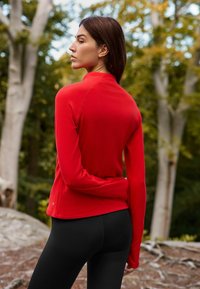 Red long-sleeve top with a high collar, fitted design, and smooth texture, paired with black leggings in an outdoor setting.