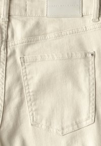 Tissu en denim beige avec une poche arrière de forme carrée et des coutures subtiles. Comprend un petit détail de bouton sur la poche.