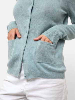 Cardigan azzurro chiaro realizzato in tessuto morbido, con chiusura a bottoni, polsini a coste e due tasche frontali. La texture appare liscia.