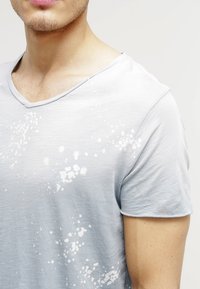 Camiseta de algodón azul claro con escote en V, que presenta un sutil patrón de salpicaduras en blanco. Textura suave y mangas cortas.
