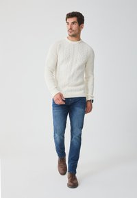 Pulover alb tricotat cu model din cabluri, asociat cu blugi albaștri și cizme din piele maro. Modelul stă pe un fundal simplu, deschis la culoare.